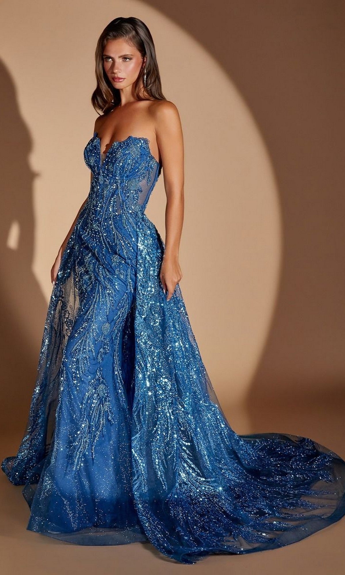A woman wearing the Ladivine CM393 long prom dress in blue—a floor-length sequined gown featuring a strapless, sheer bodice and a flowing train—stands against a neutral background.